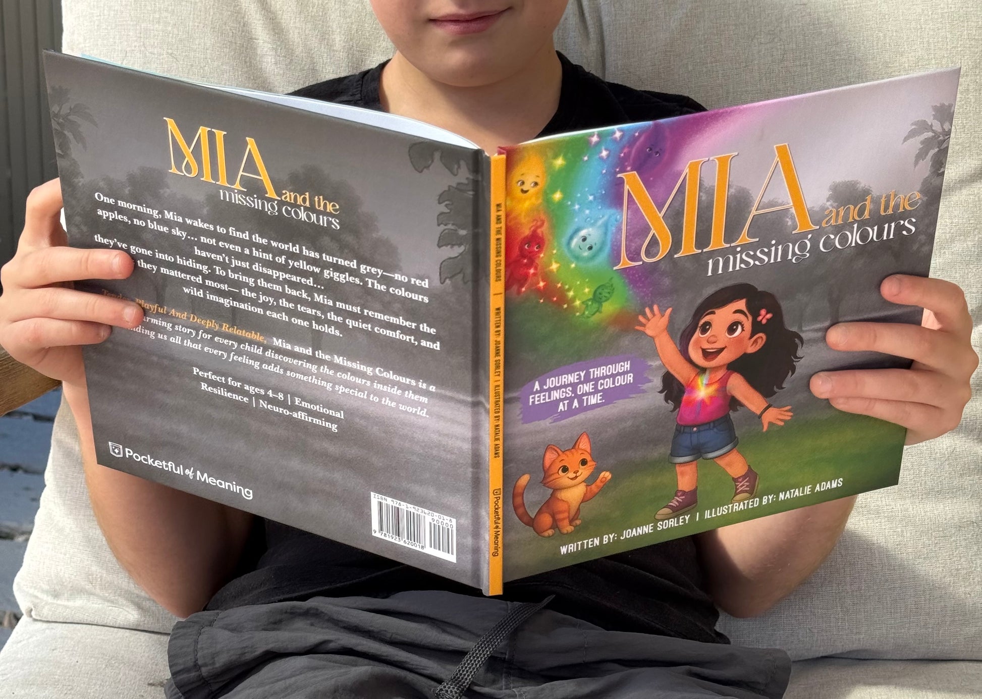 Child holding a book titled 'Mia and the Missing Colours' with colorful cover design.